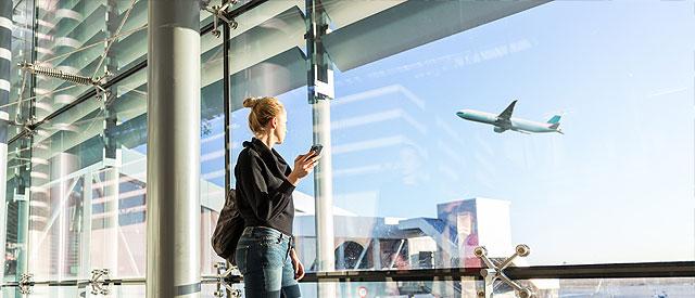 a woman standing in an airport looking out the window at a plane in the sky.